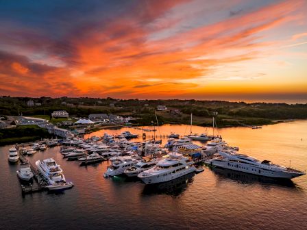 A vibrant sunset over a marina with numerous yachts and boats docked. The sky has shades of orange and pink, reflecting on the calm water.