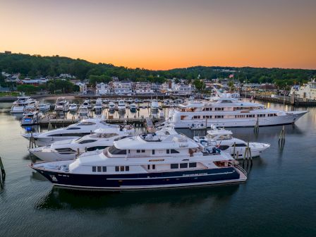 A marina filled with luxury yachts at sunset, with a hilly landscape and buildings in the background.