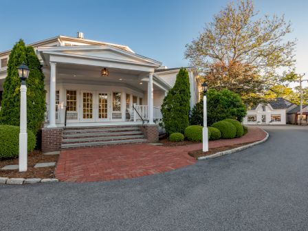 A large white building with columns, shrubs, and a brick path with a road curving around it. It appears to be a residential or public facility.
