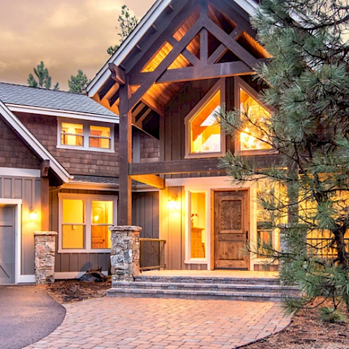 A well-lit, modern wooden house with large windows, a double garage, and a stone pathway, surrounded by tall trees.
