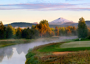 A scenic view showcasing a golf course alongside a tranquil river, surrounded by trees, with a snow-capped mountain in the background.