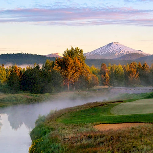 A scenic view showcasing a golf course alongside a tranquil river, surrounded by trees, with a snow-capped mountain in the background.