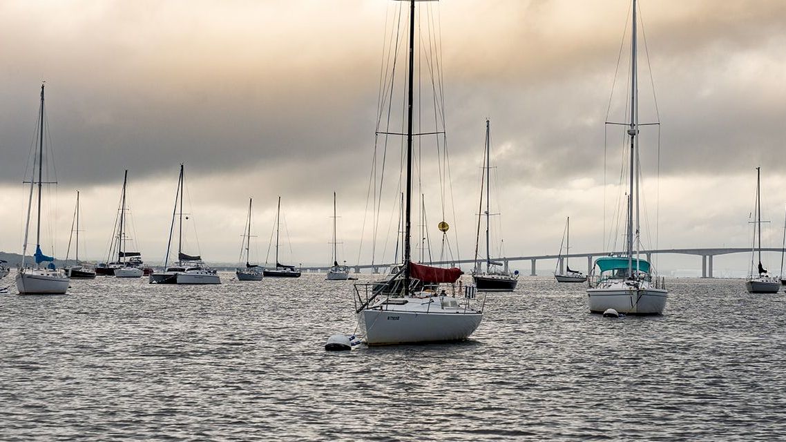 Several sailboats are anchored on calm water under a cloudy sky, with a bridge in the background at the horizon, creating a peaceful scene.