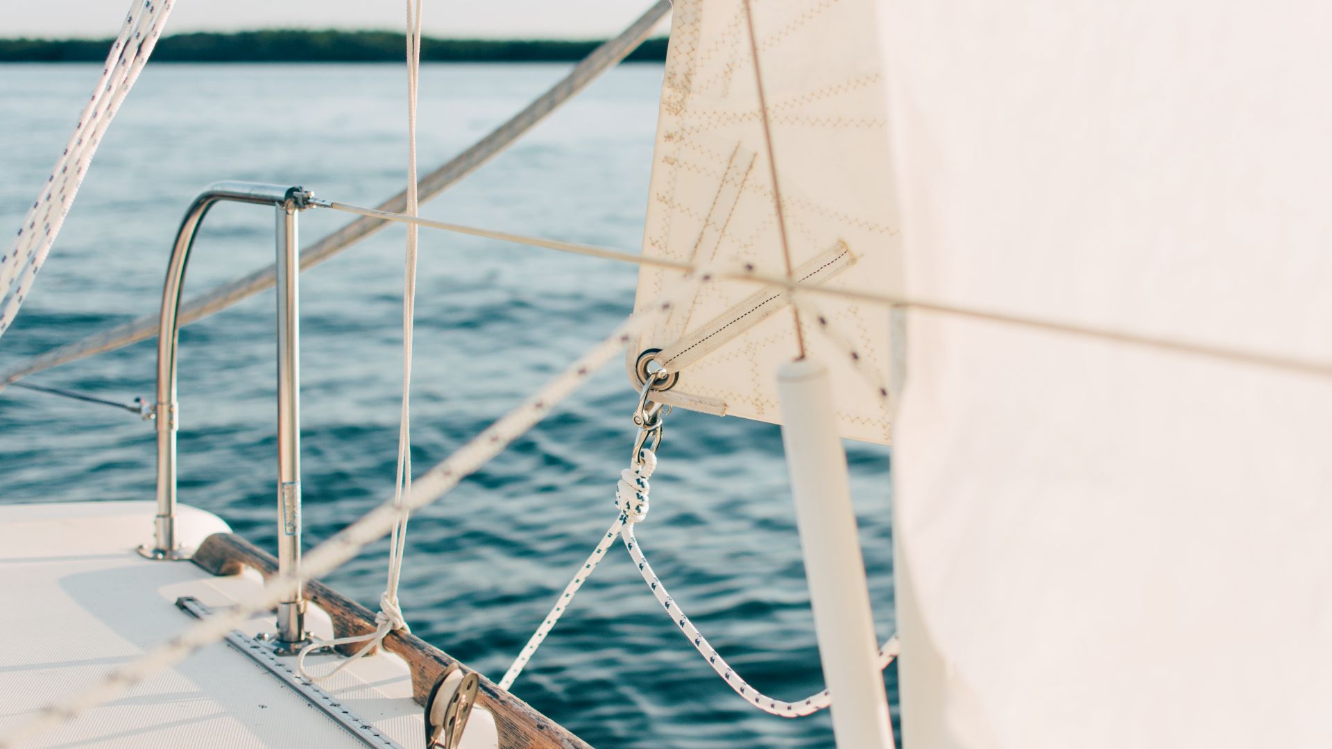Close-up of a sailboat's deck, showing a metal railing, white sail, and various ropes and chains, with the blue water in the background.